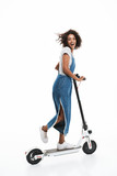 Image of excited african american woman dressed in denim overalls smiling at camera while riding on scooter