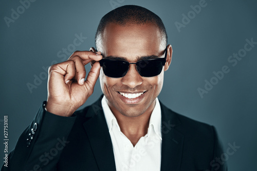 Everything must be perfect. Charming young African man in full suit looking at camera and adjusting eyewear while standing against grey background photo