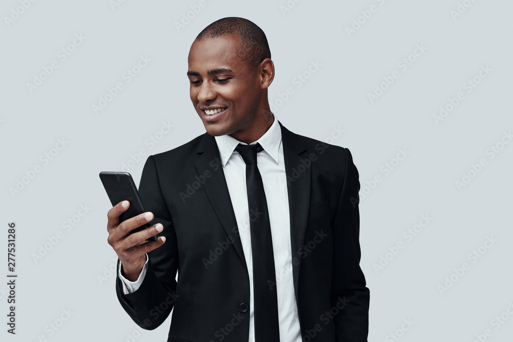 Text messaging. Charming young African man in formalwear using smart phone and smiling while standing against grey background Text messaging. Charming young African man in formalwear using smart phone and smiling while standing against grey background