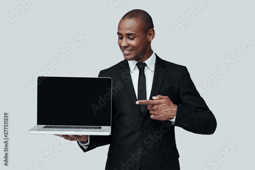 Just look here! Young African man in formalwear pointing at laptop and smiling while standing against grey background