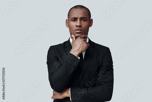 Time to make decision. Handsome young African man in formalwear looking at camera and keeping hand on chin while standing against grey background