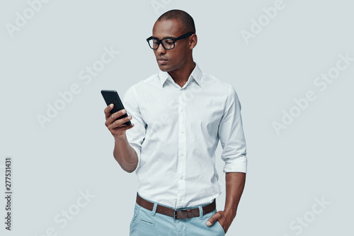 Very busy. Handsome young African man using smart phone while standing against grey background © G-Stock Faces