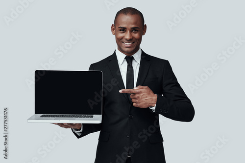 You should see this! Young African man in formalwear pointing at laptop and smiling while standing against grey background