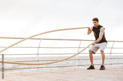 Photo of unshaven man in tracksuit working out with fitness rope on wooden pier at seaside © Drobot Dean