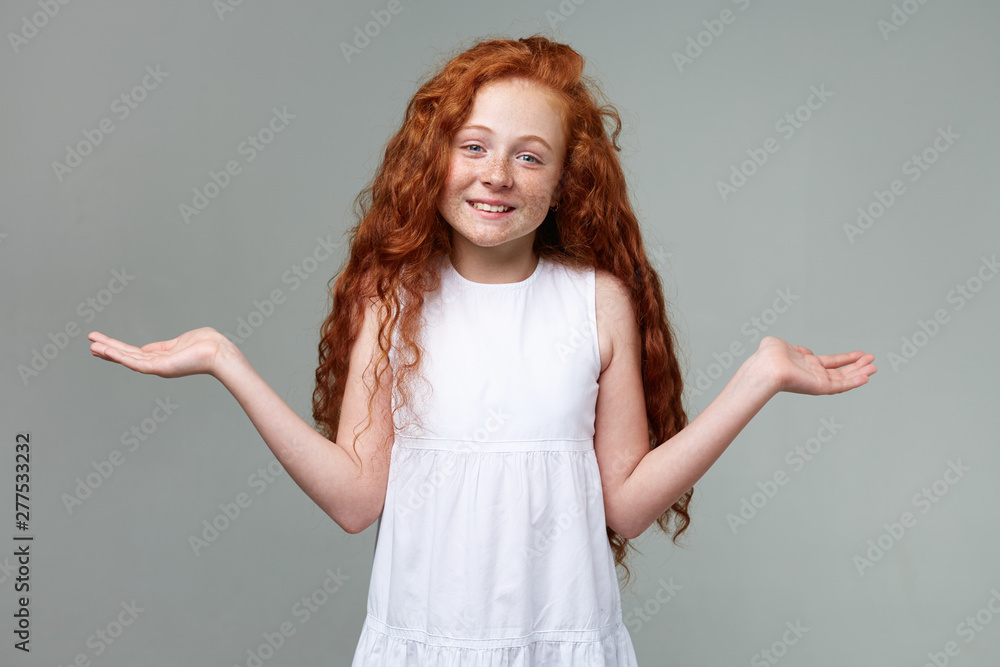 Portrait of distracted nice little girl with ginger hair and freckles, looks confused, spreads his hands to the side, stands over gray background. Portrait of distracted nice little girl with ginger hair and freckles, looks confused, spreads his hands to the side, stands over gray background.