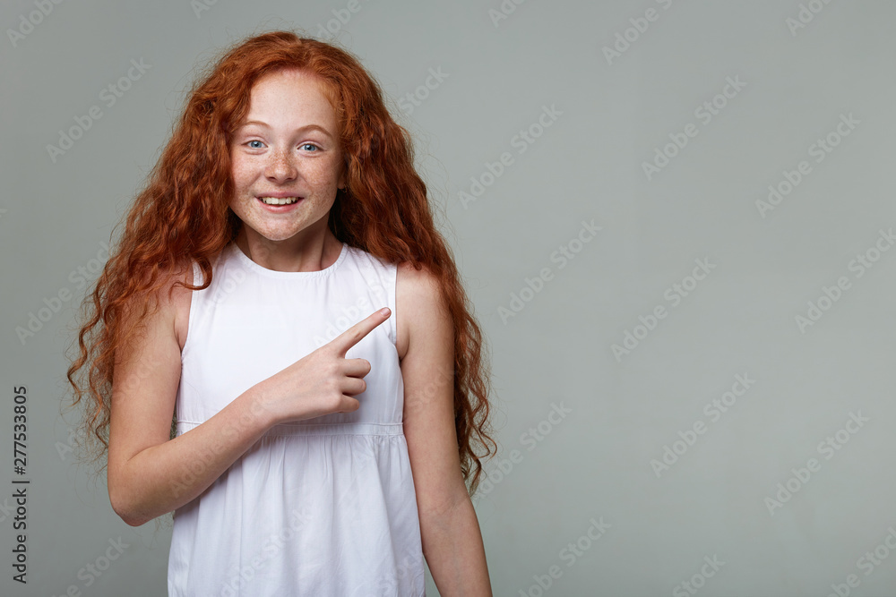 Nice happy little girl with ginger hair and freckles, smiles and looks at the camera, wonnts to draw you attention and points fingers to the copy space on the right side, stands over gray background . Nice happy little girl with ginger hair and freckles, smiles and looks at the camera, wonnts to draw you attention and points fingers to the copy space on the right side, stands over gray background .