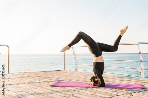 Beautiful young brunette woman in a black jumpsuit practicing yoga on the beach at sunrise. Concept of wellness and healthy lifestyle.