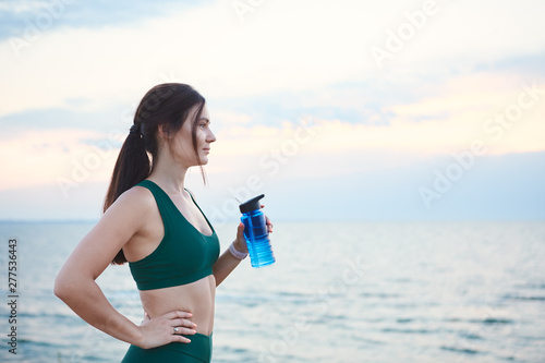 Young brunette woman with wireless earbuds and smart watches resting after morning workout drinking water.