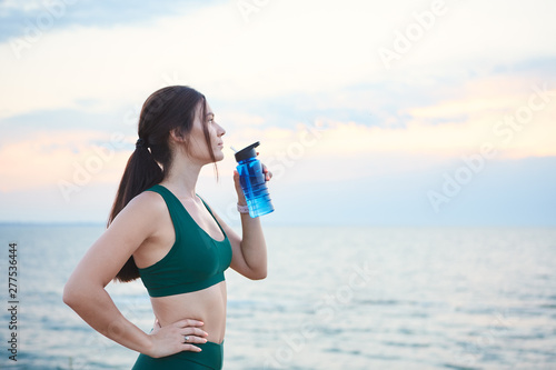 Young brunette woman with wireless earbuds and smart watches resting after morning workout drinking water.