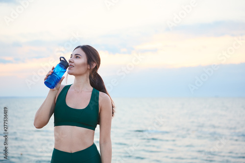 Young brunette woman with wireless earbuds and smart watches resting after morning workout drinking water.