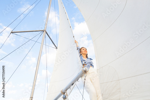 A girl with long hair, a model sits on a yacht, in white pants, a blue shirt, sunglasses, beside a sail, against a blue sky with space for an inscription. The concept of vacation at sea.