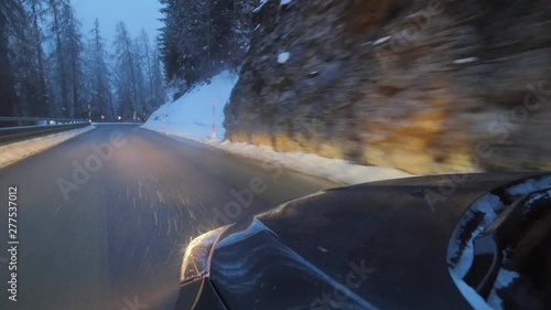 Black automibile with headlights on is smoothly and carefully moving on road in mountain area along snow covered forest trees and hills, in the cold winter evening. photo