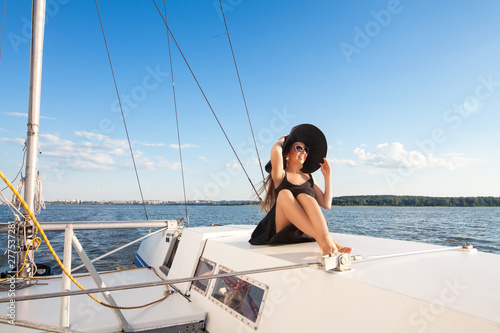A girl model, brunette in a black board, with long hair, wearing a hat, wearing sunglasses, sits on a white yacht, next to a sail, against a blue clear sky, blue sea, with space for an inscription. 