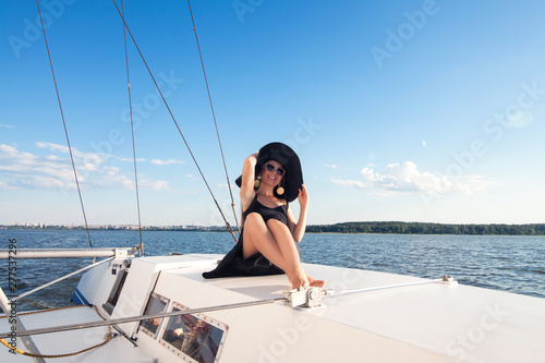 A girl model, brunette in a black board, with long hair, wearing a hat, wearing sunglasses, sits on a white yacht, next to a sail, against a blue clear sky, blue sea, with space for an inscription.  © Карина Желнина