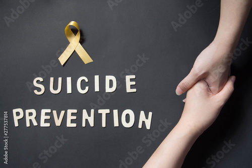 World suicide prevention day - Holding hands for helping and supporting depressed woman with yellow ribbon awareness and SUICIDE PREVENTION wooden word on black background. Mental health care concept.