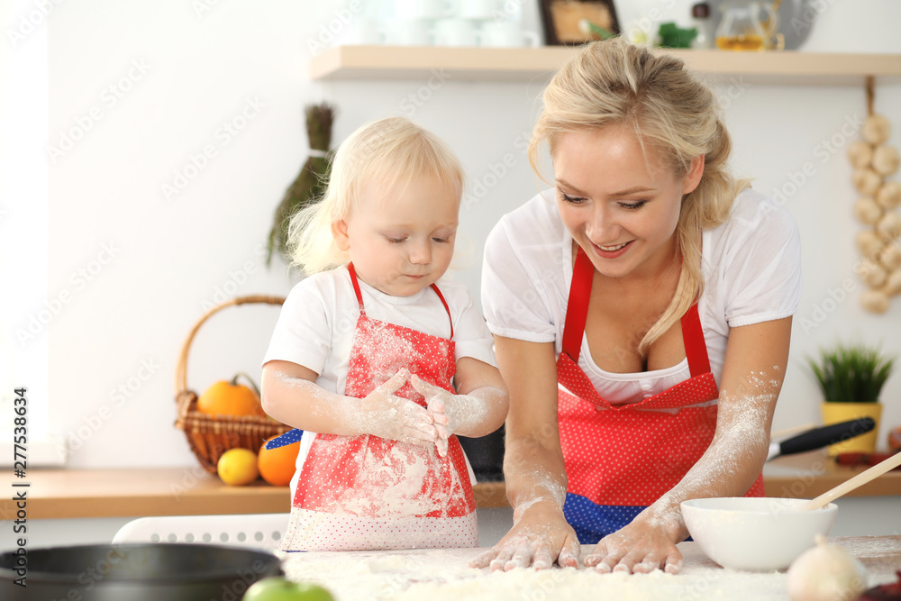 Little girl and her blonde mom in red aprons playing and laughing while kneading the dough in kitchen. Homemade pastry for bread, pizza or bake cookies. Family fun and cooking concept
