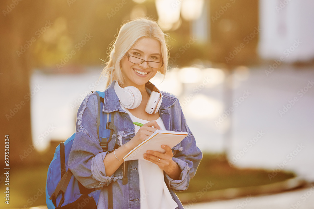 Portrait of a smiling young woman student in eyeglasses with backpack listening music with headphones and writing notes. Online and remote education concept