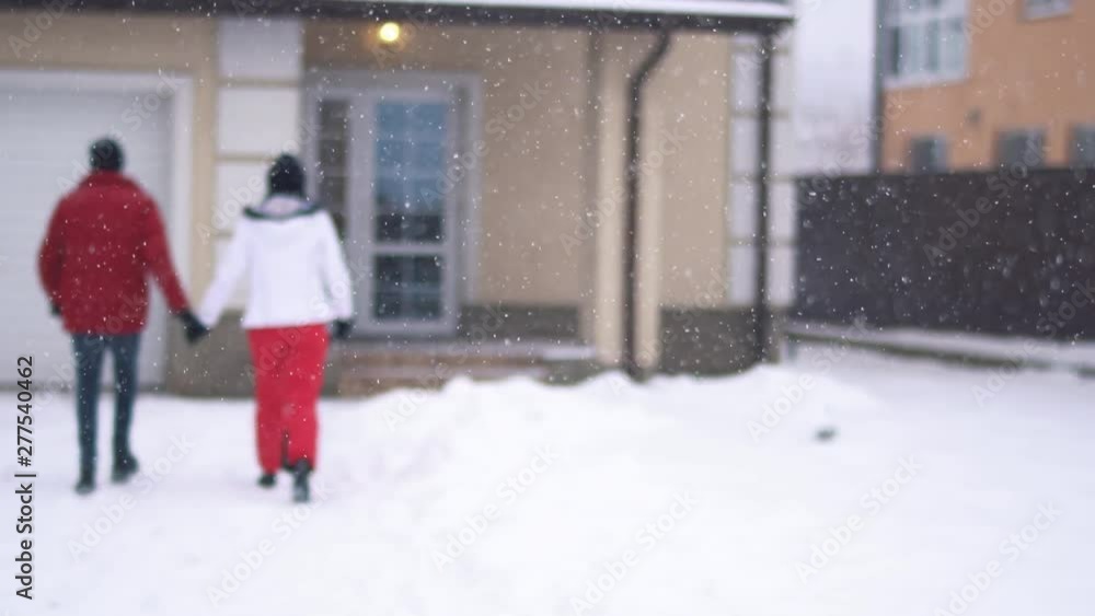 Young woman and bearded man walking on the backyard toward house, holding hands. Happy couple have a fun in winter backyard. Snow is falling. Concept of outdoor recreation. Back shooting Young woman and bearded man walking on the backyard toward house, holding hands. Happy couple have a fun in winter backyard. Snow is falling. Concept of outdoor recreation. Back shooting