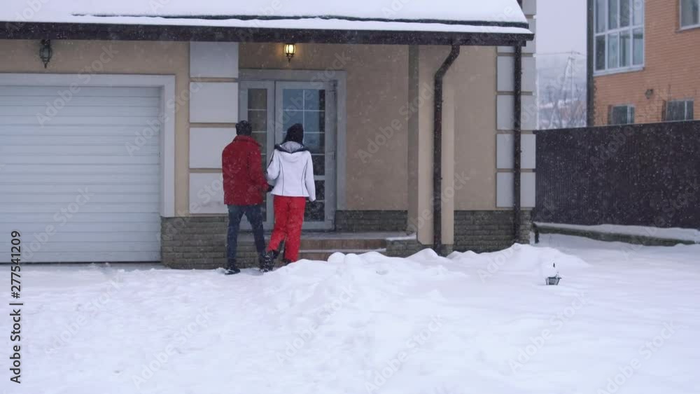 Young woman and bearded man walking on the backyard toward house, holding hands. Happy couple have a fun in winter backyard. Snow is falling. Concept of outdoor recreation. Back shooting Young woman and bearded man walking on the backyard toward house, holding hands. Happy couple have a fun in winter backyard. Snow is falling. Concept of outdoor recreation. Back shooting