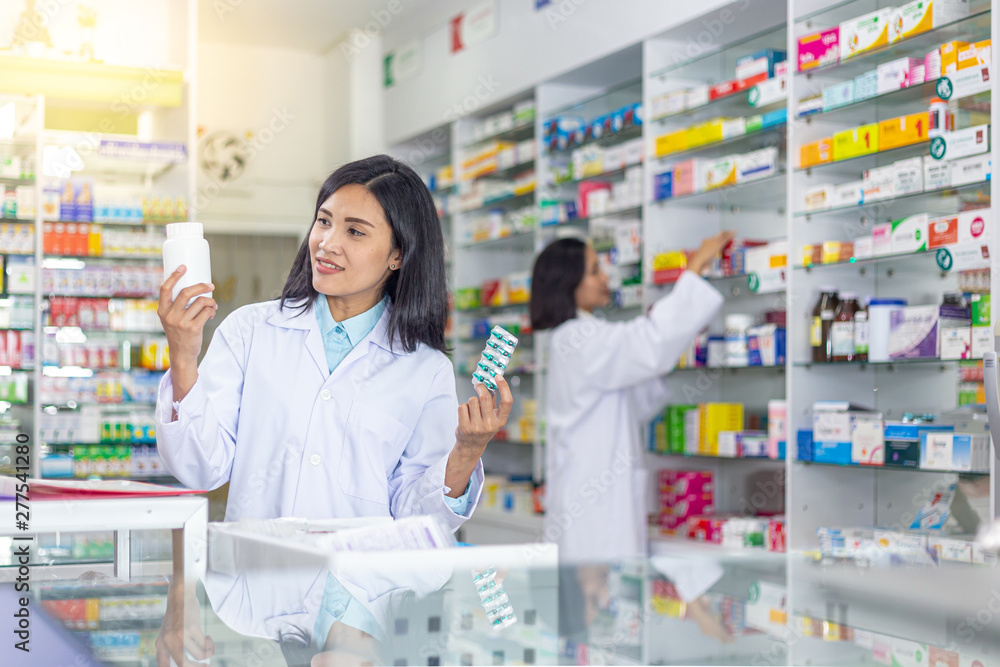 Smiling Asian female pharmacist holding medicine bottle and capsules medicine in the modern drugstore with her workmate.