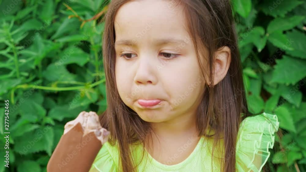 A little cute and funny girl in a bright green dress grimaces and eats chocolate ice cream. In the background is a green hedge of Virginia creeper (Parthenocissus quinquefolia). A little cute and funny girl in a bright green dress grimaces and eats chocolate ice cream. In the background is a green hedge of Virginia creeper (Parthenocissus quinquefolia).