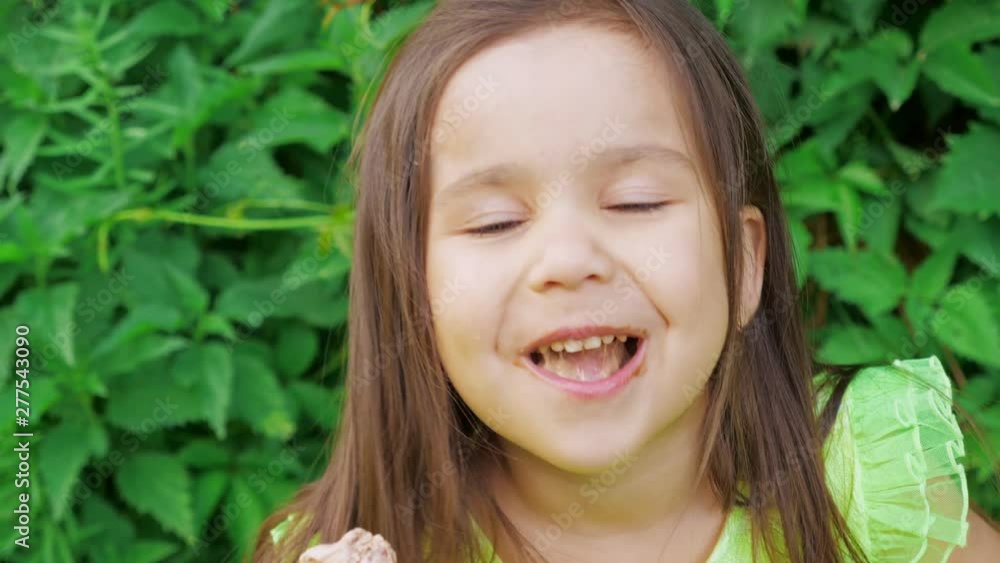 A little cute and funny girl in a bright green dress grimaces and eats chocolate ice cream. In the background is a green hedge of Virginia creeper (Parthenocissus quinquefolia). A little cute and funny girl in a bright green dress grimaces and eats chocolate ice cream. In the background is a green hedge of Virginia creeper (Parthenocissus quinquefolia).