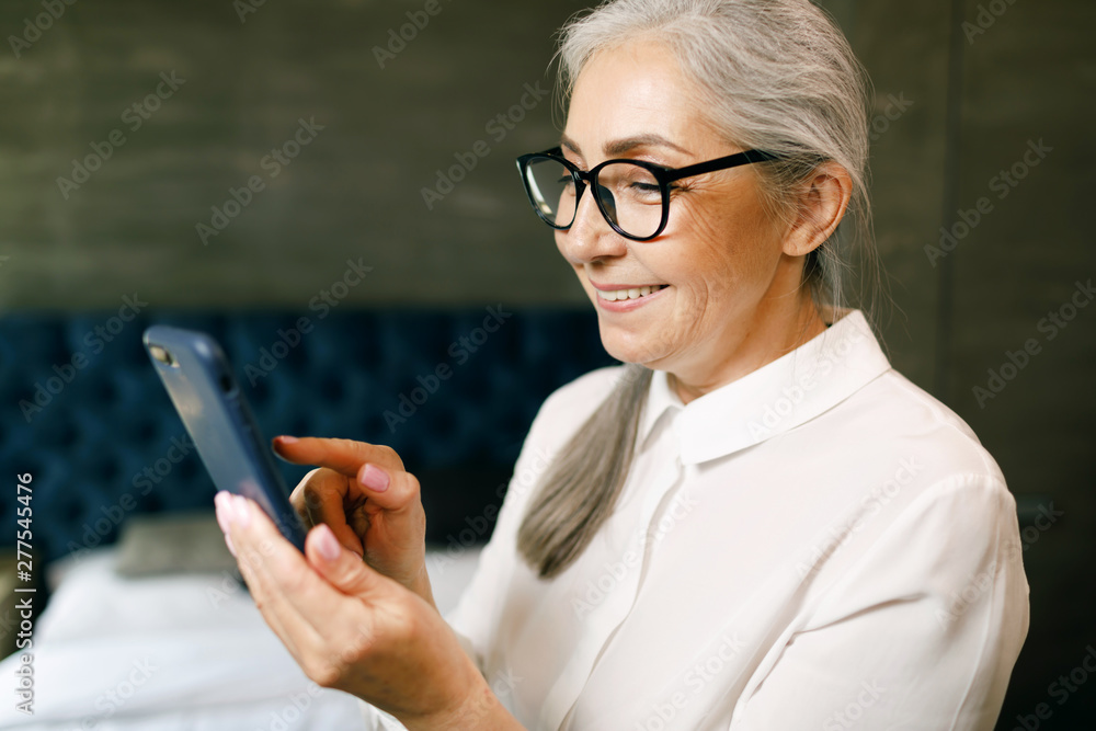 Senior woman with grey hair in eyeglasses typing message on smartphone