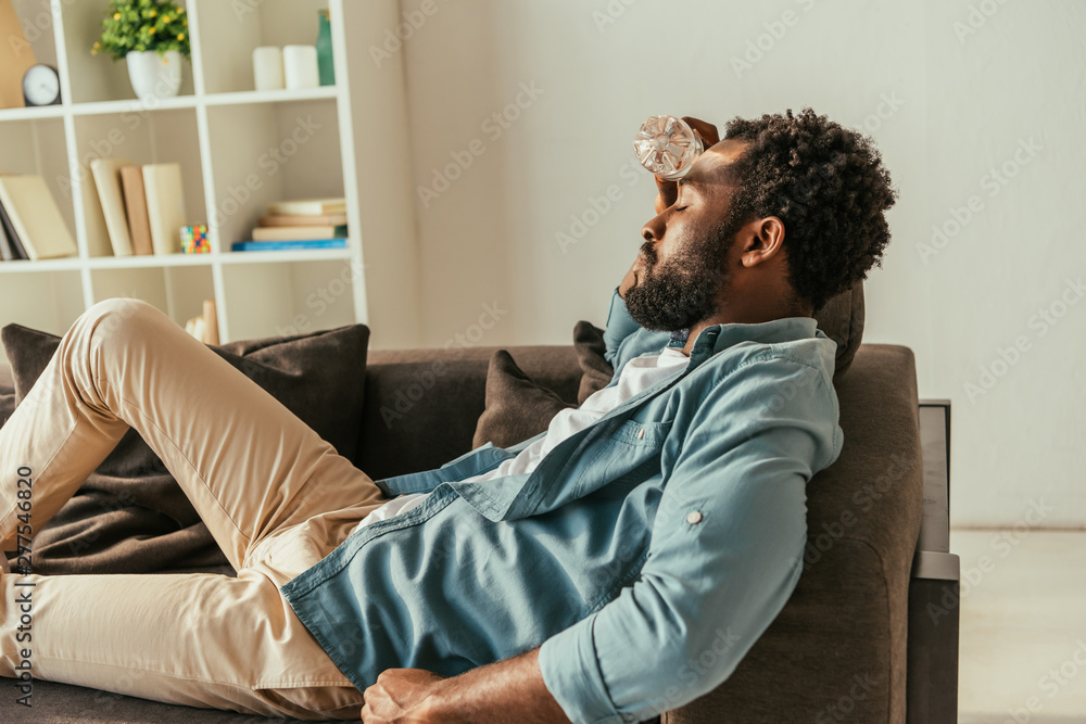 exhausted african american man lying on couch with closed eyes and holding bottle with water near head while suffering from heat exhausted african american man lying on couch with closed eyes and holding bottle with water near head while suffering from heat