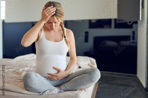 Troubled heavily pregnant young woman sitting cross legged on a bed cradling her swollen abdomen and clutching her head looking down with a serious expression