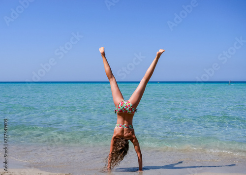 Slim and athletic girl doing a handstand before a wonderful beach with crystal clear water -vacation-fitness-wellness