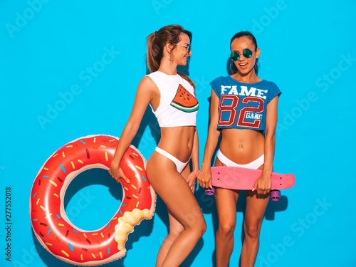 Two beautiful sexy smiling hipster women in summer underpants and topic.Girls in sunglasses. Positive models having fun with colorful penny skateboards.With donut lilo inflatable mattress