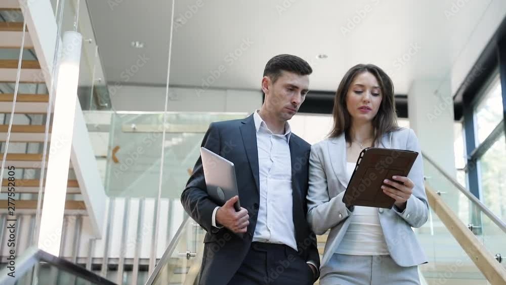 Two young Successful male and female Scientist walking in big bright Building. Collegues talking and using New Gadgets: Tablet, Laptop for checking News. Nanotechnology. Teamwork. Partnership. App. Two young Successful male and female Scientist walking in big bright Building. Collegues talking and using New Gadgets: Tablet, Laptop for checking News. Nanotechnology. Teamwork. Partnership. App.