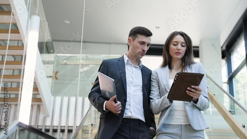 Two young Successful male and female Scientist walking in big bright Building. Collegues talking and using New Gadgets: Tablet, Laptop for checking News. Nanotechnology. Teamwork. Partnership. App.