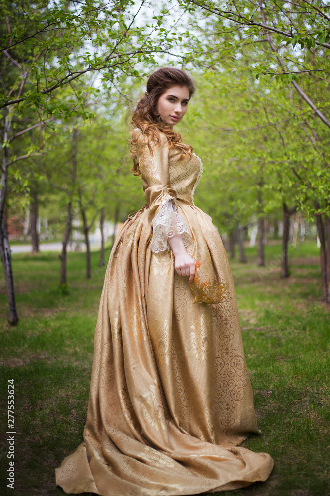 Beautiful girl in a gold dress against a background of green trees. A woman in a ball gown of the Rococo era. Beautiful girl in a gold dress against a background of green trees. A woman in a ball gown of the Rococo era.