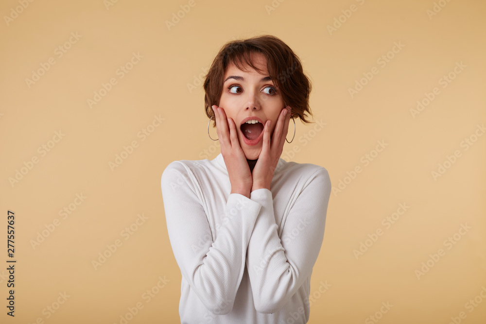 Cute happy amazed short haired young woman in blank t-shirt, loks surprised with wide open mouth, screaming and looks away, touches cheeks, stands over beige wall. Cute happy amazed short haired young woman in blank t-shirt, loks surprised with wide open mouth, screaming and looks away, touches cheeks, stands over beige wall.