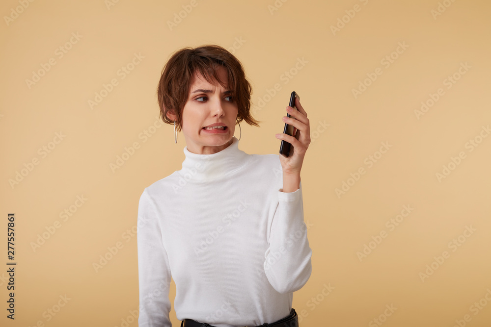 Portrait of short haired young woman wears in white golf, keeps the phone away from your ear, looks at the display with disgusted expression, looks shocked, stands over beige background. Portrait of short haired young woman wears in white golf, keeps the phone away from your ear, looks at the display with disgusted expression, looks shocked, stands over beige background.