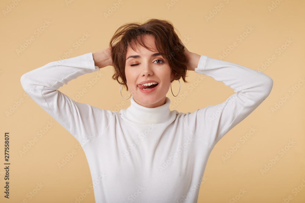 Playful short haired young woman wears in white golf, holds his head with arms, looks at the camera wit funny expression, looks happy, stands over beige background and winks. Playful short haired young woman wears in white golf, holds his head with arms, looks at the camera wit funny expression, looks happy, stands over beige background and winks.
