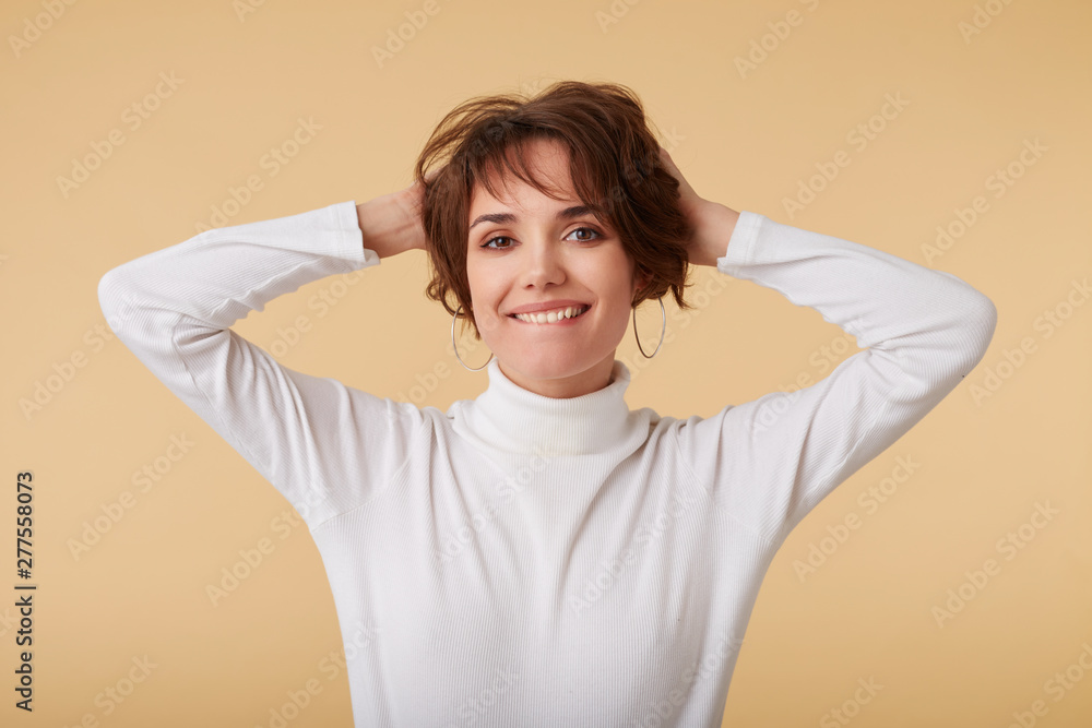 Joyful short haired young woman wears in white golf, holds his head with arms, looks at the camera wit funny expression, looks calm, stands over beige background. Joyful short haired young woman wears in white golf, holds his head with arms, looks at the camera wit funny expression, looks calm, stands over beige background.