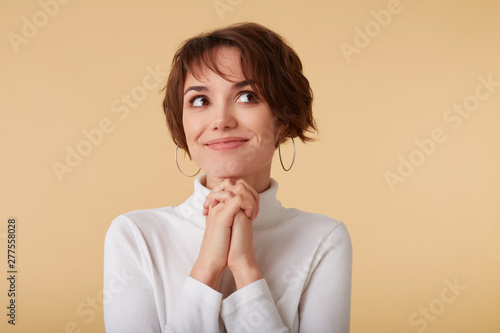 Close up of thinking short haired young woman wears in white golf, stands over beige background, dreamily looks away.