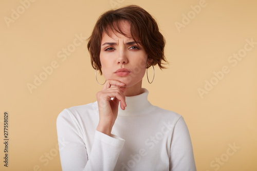 Close up of doubtful short haired young woman wears in white golf, stands over beige background, pensive looks at the camera and touches chin.