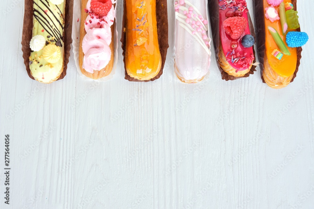 Fototapeta Set of design delicious eclairs with creative colorful decor on white textured table. Tasty French eclairs with icing, cream, fresh berries and sugar elements. Sweet flat lay. Homemade profiteroles