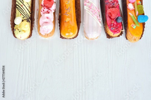Set of design delicious eclairs with creative colorful decor on white textured table. Tasty French eclairs with icing, cream, fresh berries and sugar elements. Sweet flat lay. Homemade profiteroles