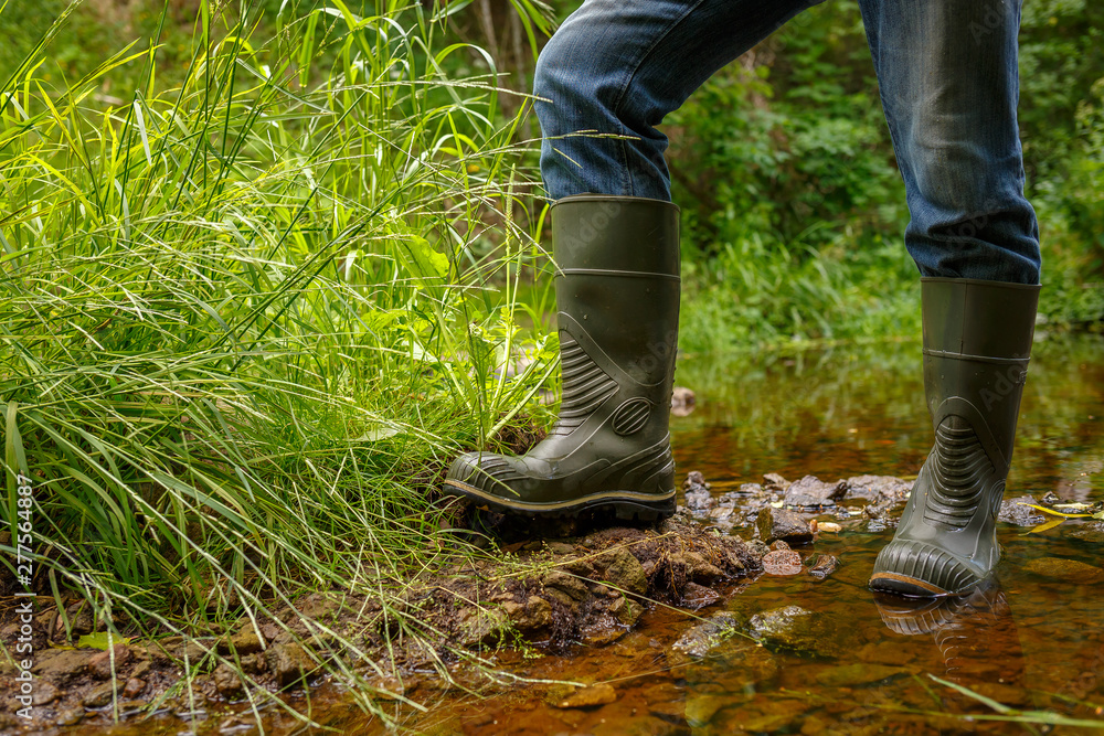 A man in rubber boots is standing on the bank of a forest stream