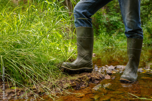 A man in rubber boots is standing on the bank of a forest stream. Crossing over the creek. Waterproof footwear for outdoor activities, fishing, hunting and hiking. Reliable shoes for country walks.