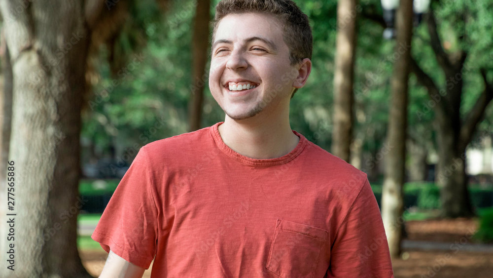 A young man with a goatees smiles very big and proudly squinting his eyes in joy looking out at the sunset wearing a bright red shirt surrounded by many trees in a beautiful city park A young man with a goatees smiles very big and proudly squinting his eyes in joy looking out at the sunset wearing a bright red shirt surrounded by many trees in a beautiful city park