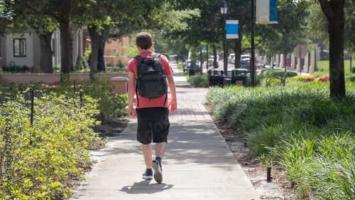 The back of young student walks hastily down the sidewalk of campus on a sunny day wearing a bright red shirt black shorts and blue athletic shoes with his black and grey backpack hanging on his back