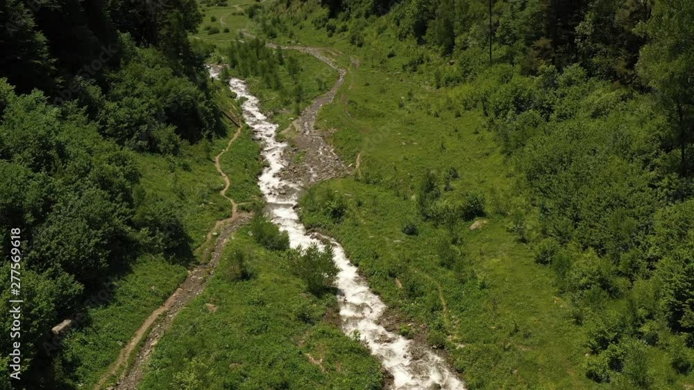 Winding mountain stream flowing along hiking trail and pine forested mountain. Charming village located in picturesque valley. Epic mountain ranges with snowy peaks against blue sky Winding mountain stream flowing along hiking trail and pine forested mountain. Charming village located in picturesque valley. Epic mountain ranges with snowy peaks against blue sky