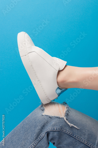 Trendy white eco leather sheakers on bent leg knee in ragged jeans over pastel turquoise blue background. Colorful main trend backdrop for your magazine, online store and media. Copy space