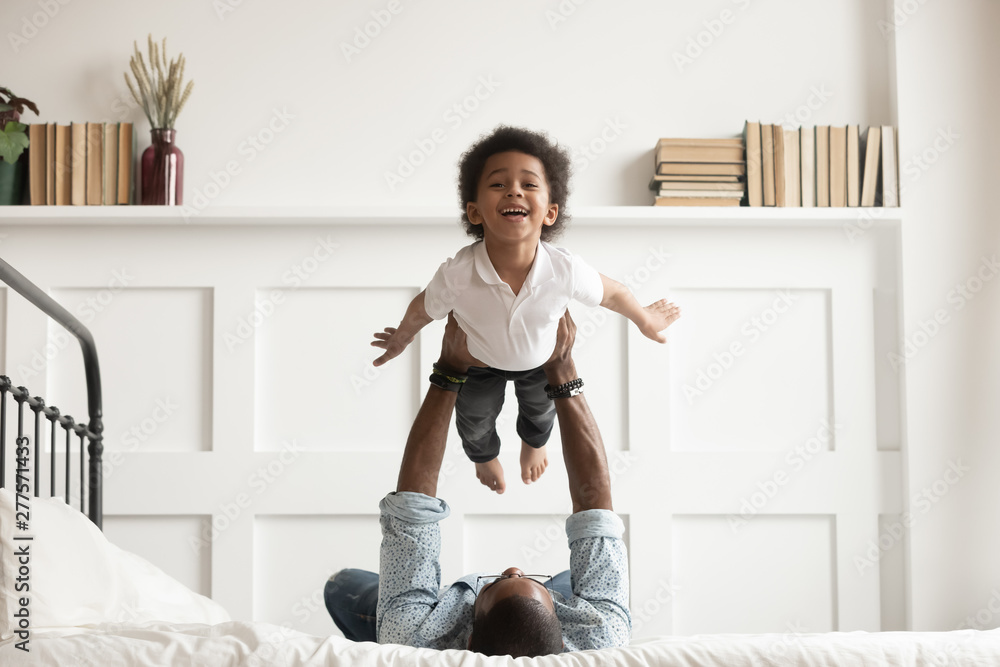 Happy african kid boy flying in fathers arms on bed - obrazy, fototapety, plakaty 