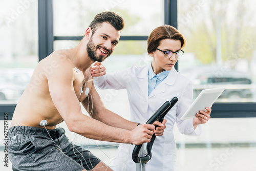 Εκτύπωση καμβά doctor with digital tablet near smiling shirtless sportsman during endurance tes
